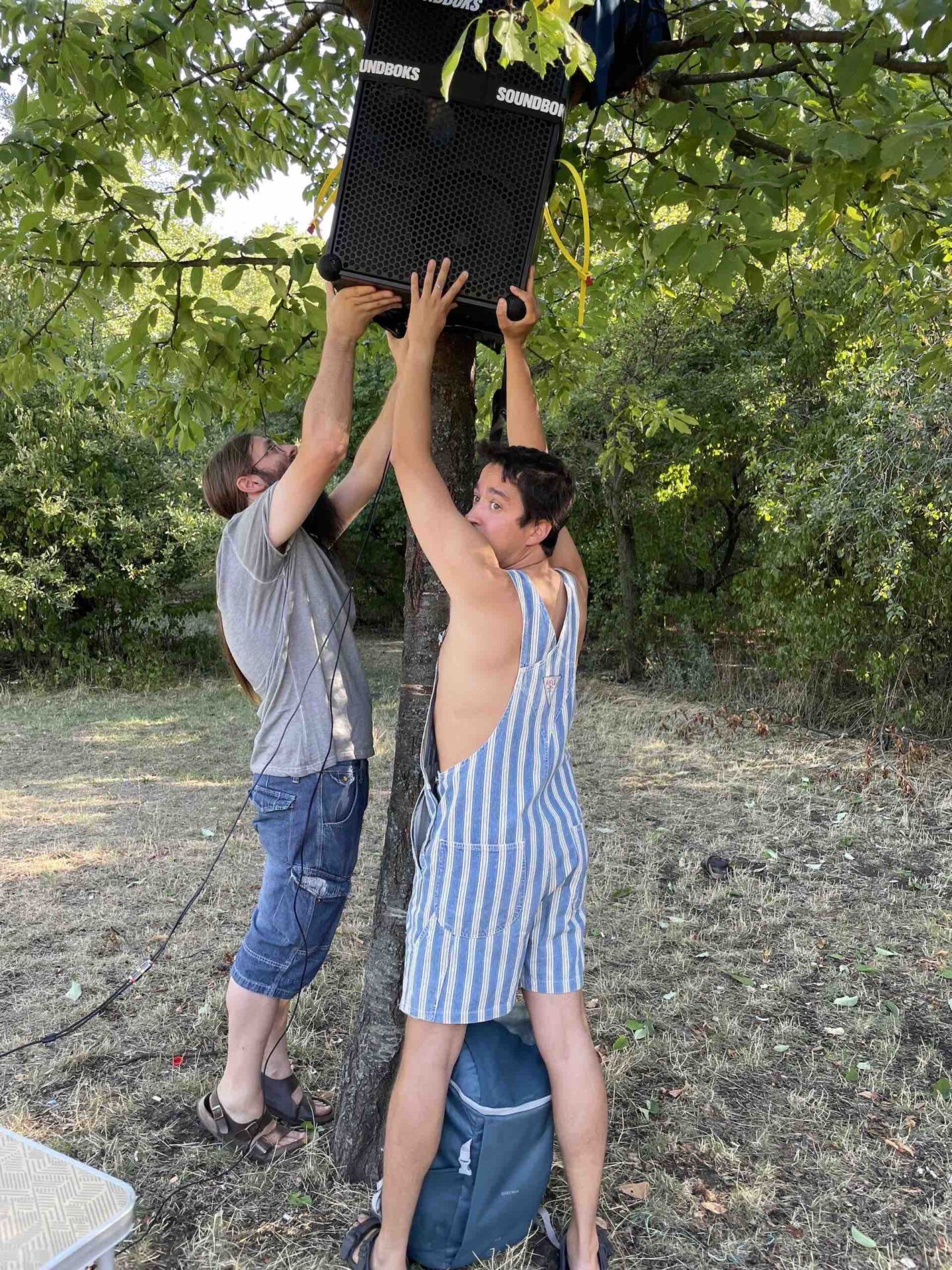 Two people preparing a rave hang a loudspeaker from a tree.